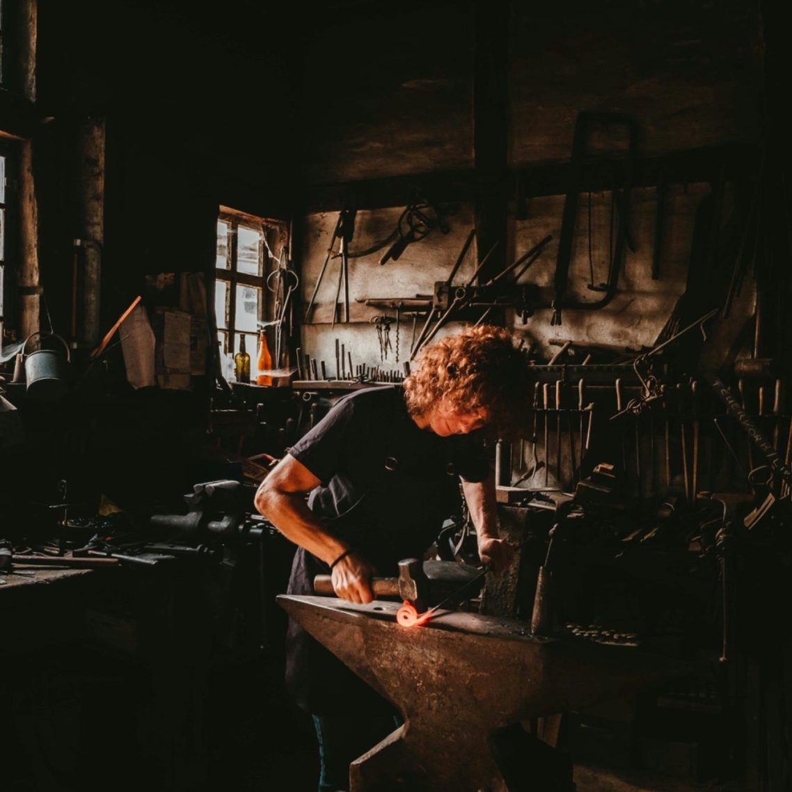 Schmied arbeitet mit glühendem Metall am Amboss in einer dunklen Werkstatt.