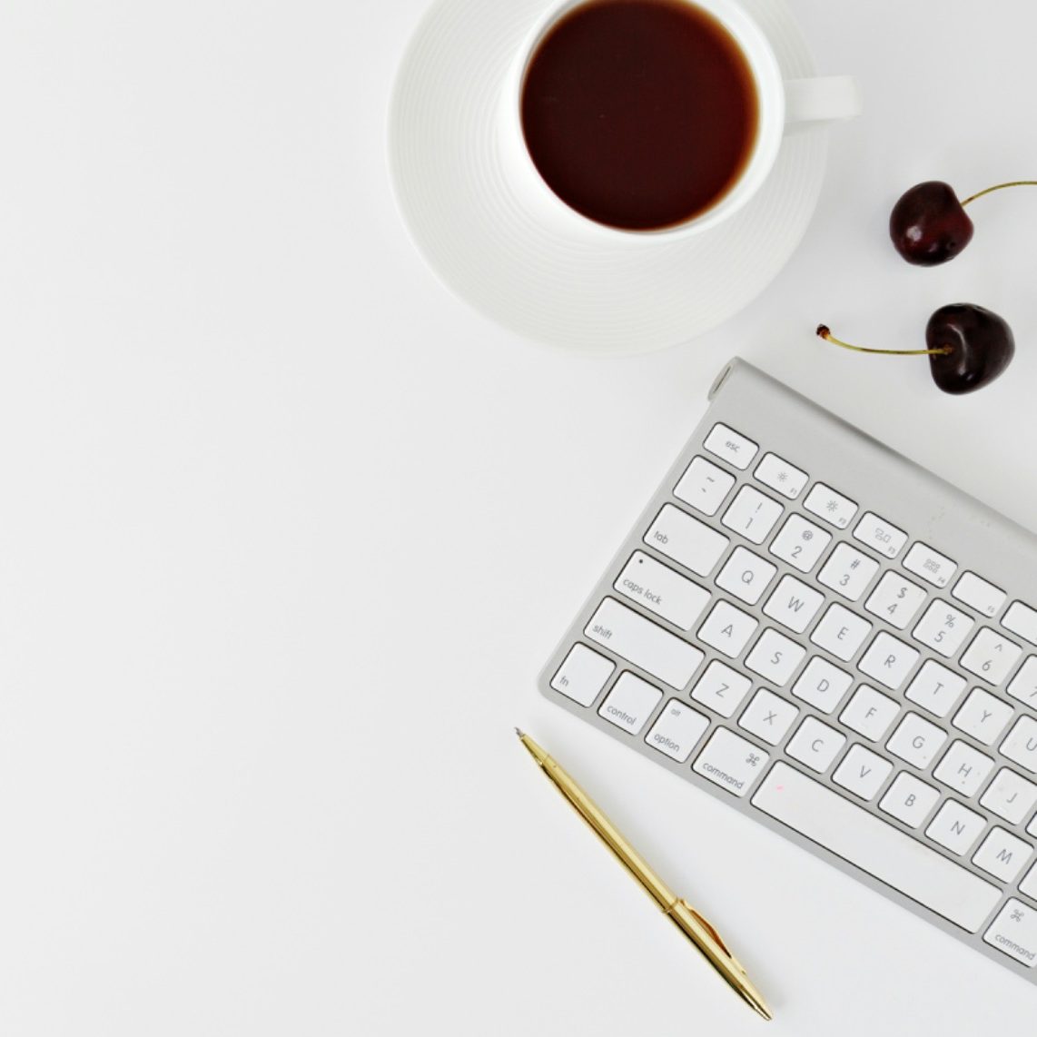 Tastatur, Goldstift, Tasse Kaffee und Kirschen auf einem weißen Tisch.